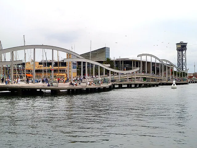 The wavy wooden Rambla de Mar footbridge crossing Port Vell, with the Maremagnum building and the cable car tower beyond