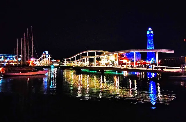 The Rambla de Mar footbridge and the lit-up Jaume I tower reflected in the water of Port Vell at night